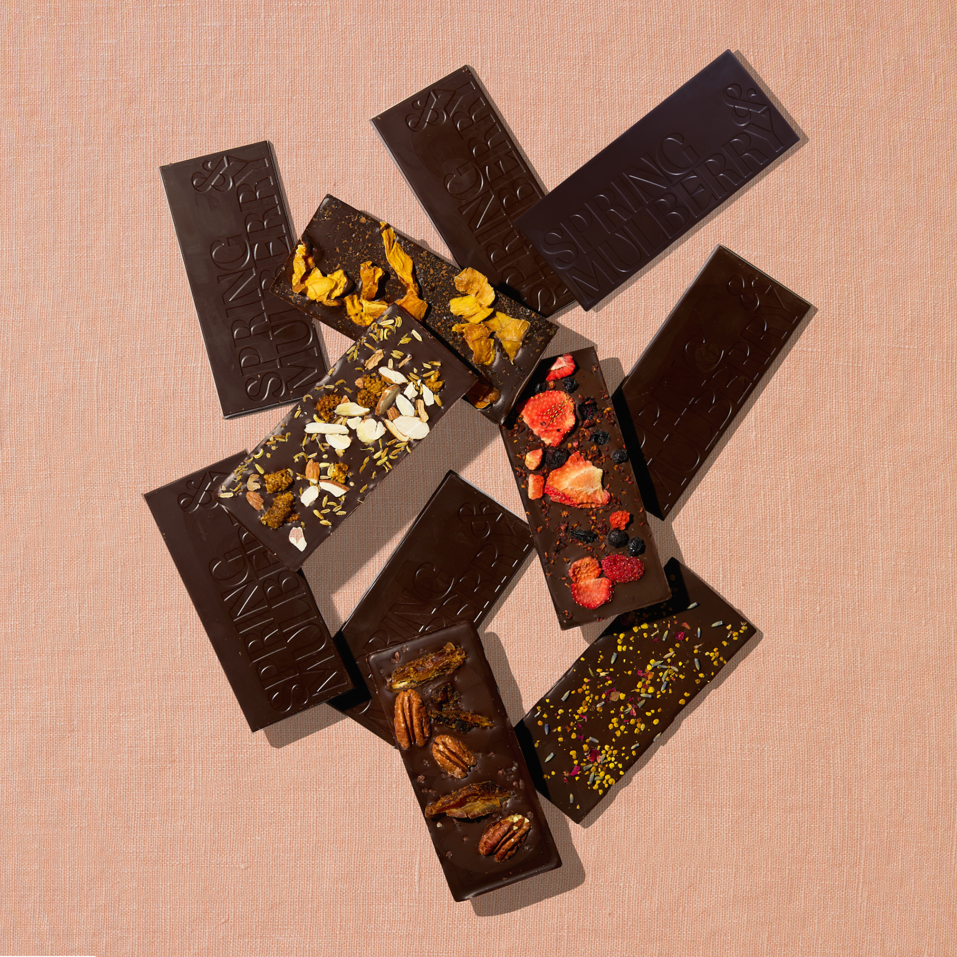 11 chocolate bars, five topped with fruits, florals, nuts, herbs and spices, and six infused with essential oils, are laid out sparadically on a light peach background.