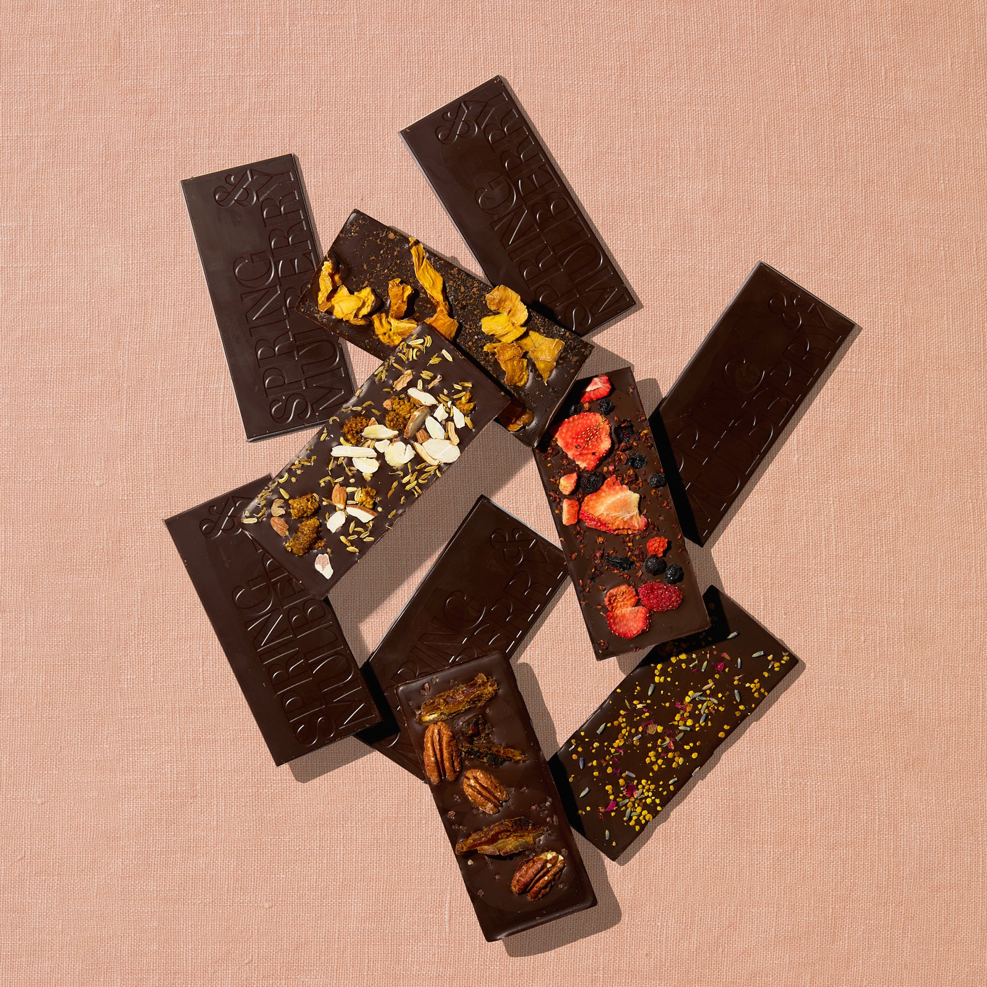 Ten chocolate bars, five topped with fruits, florals, nuts, herbs and spices, and five infused with essential oils, are laid out sparadically on a warm brown background.