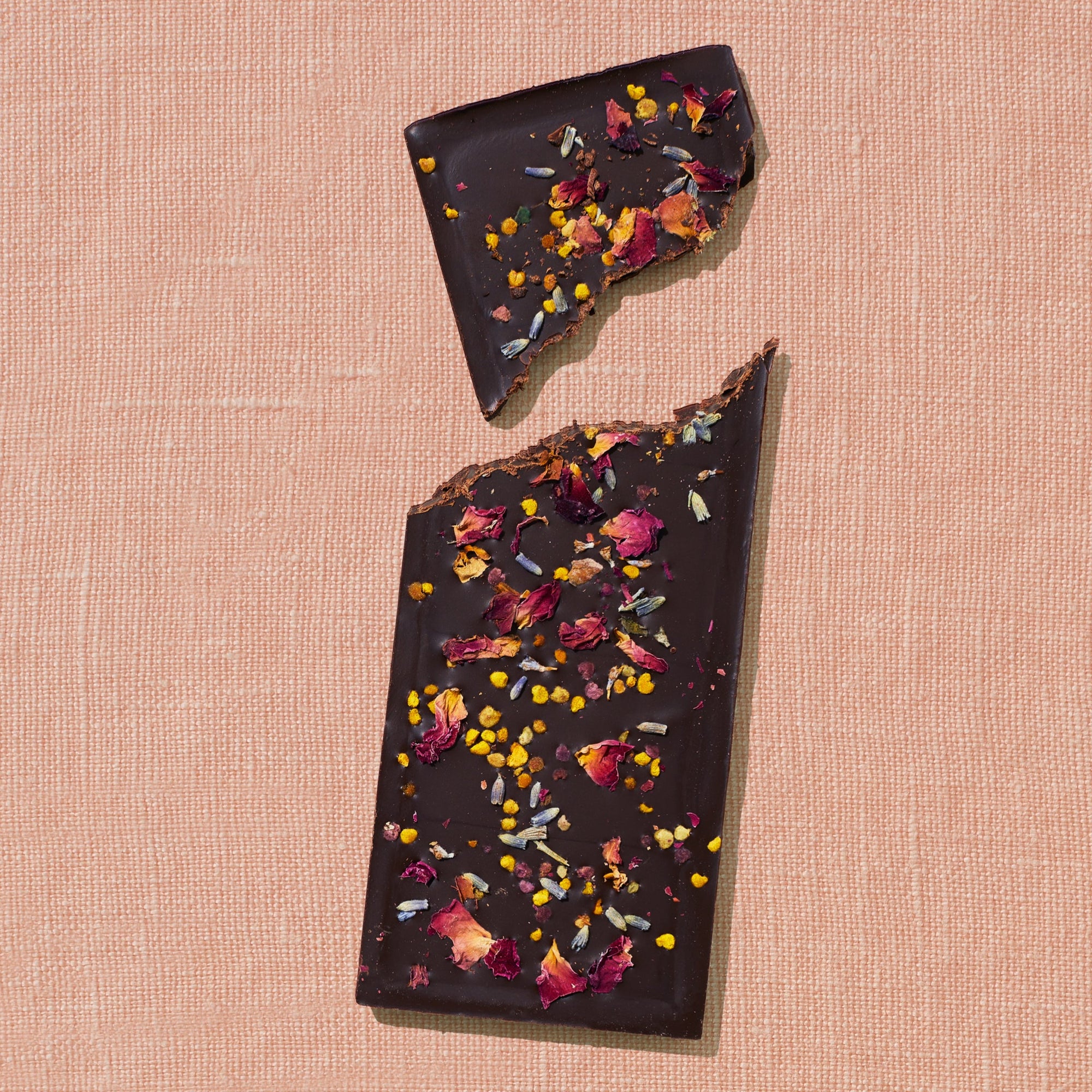 One chocolate bar that is topped with lavender, rose petals, and bee pollen is laid out on a warm brown background broken in two pieces.