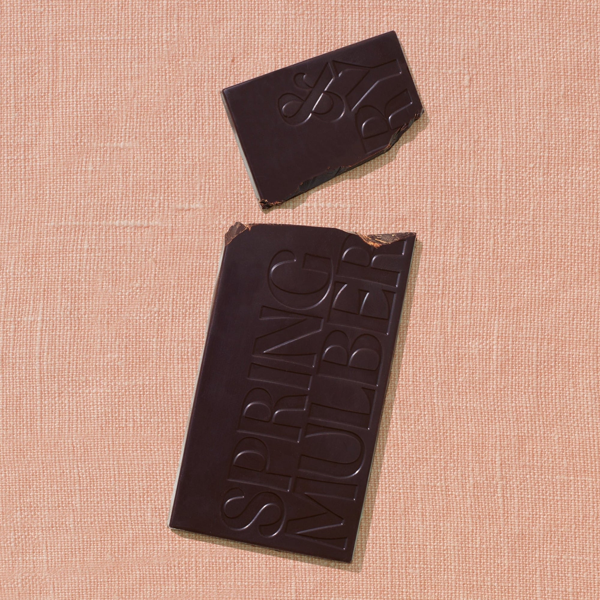 One chocolate bar that is stamped with Spring & Mulberry logo is laid out on a warm brown background broken in two pieces.