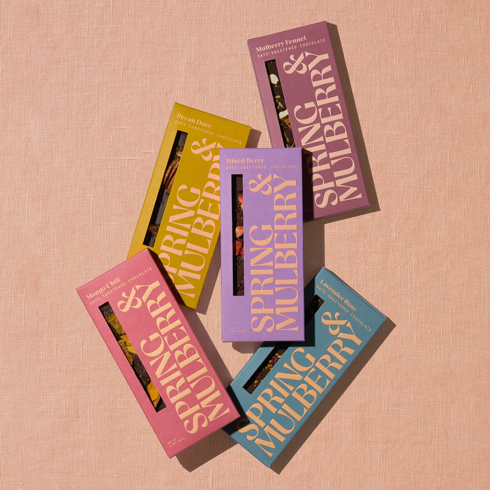 Five artisanal chocolate bars in their Spring & Mulberry boxes are laid out on a surface, each with different color packaging representing various flavors.