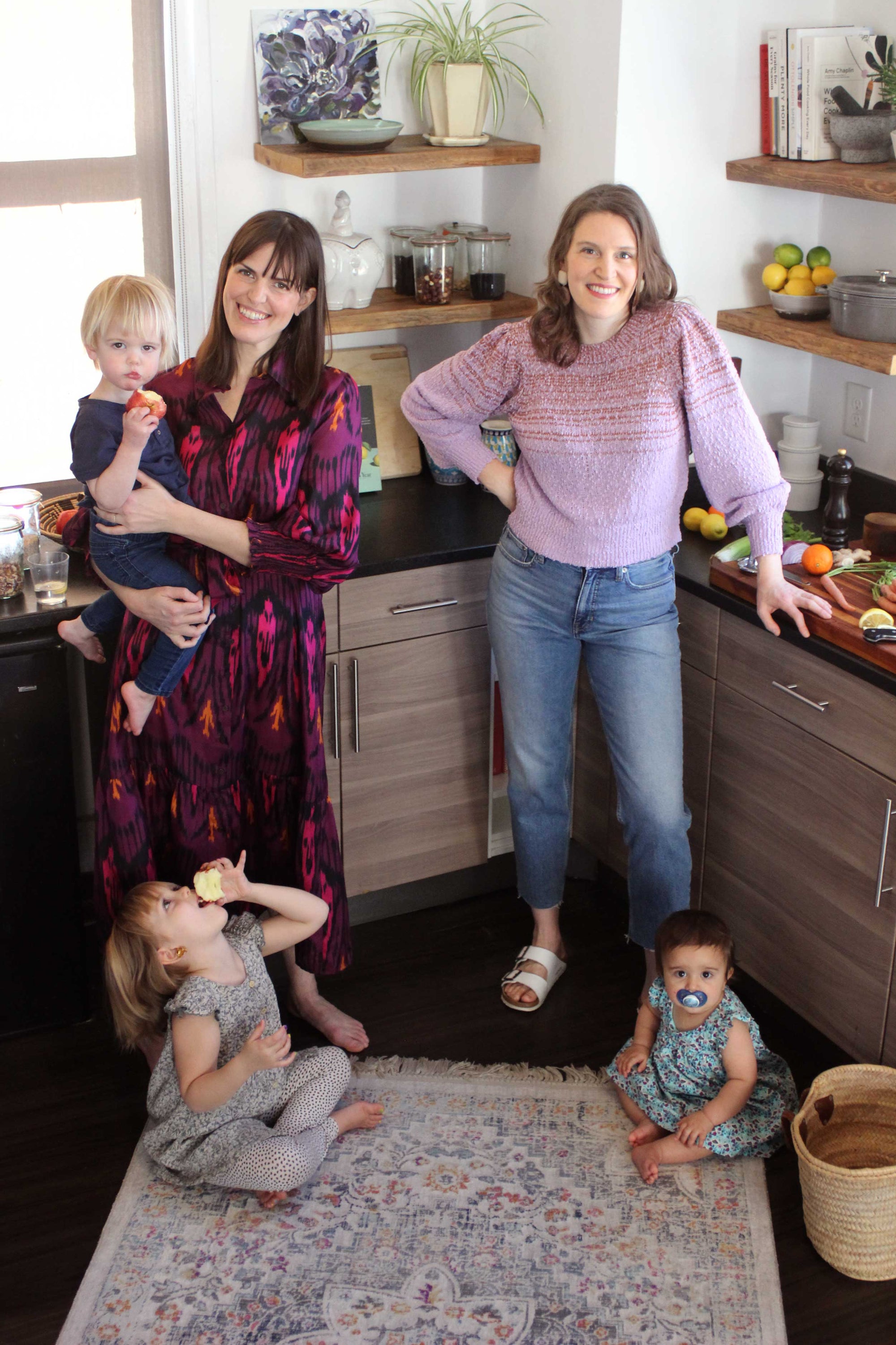 Founders Kathryn and Sarah stand in a kitchen with their three small children on the floor and in their arms. They are both tall and Caucasian with brown hair. Sarah is on the left wearing a ikat print dress Kathryn wears boyfriend jeans and a lavender sweater. She has one hand on her hip and one hand on the counter. In the background are grey-stained natural wood cabinets and stone countertops with open shelving that features cookbooks, bowls of citrus fruits, jars of beans, and a floral oil painting.