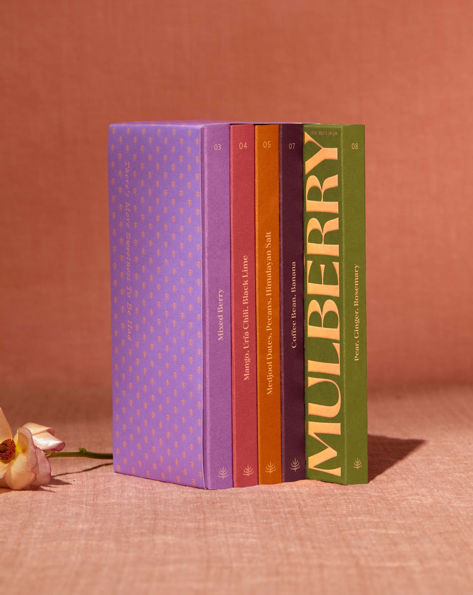 A gift set of 5 date sweetened chocolate bars with no added sugar stands on a peach linen backdrop. To the left of the gift set is a delicate flower. The gift set resembles a library set of books and is wrapped in purple paper adorned with a mulberry motif. The 5 chocolate boxes include: Mixed Berry in a purple box; Mango, Urfa Chili, Black Lime in a rose box; Medjool Date, Pecan, Himalayan Salt in a mustard box; Coffee Bean, Banana in a chocolate brown box; and, Pear, Ginger, Rosemary in a leaf green box.