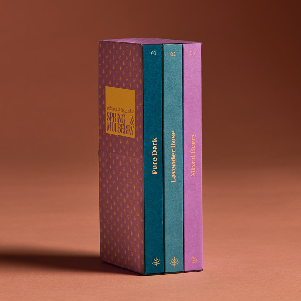A set of three artisanal chocolate bars in a vertical band, with individual bars labeled Pure Dark, Lavender Rose, and Mixed Berry, indicating different flavors. The band is lavender with a peach mulberry motif and Spring & Mulberry logo.