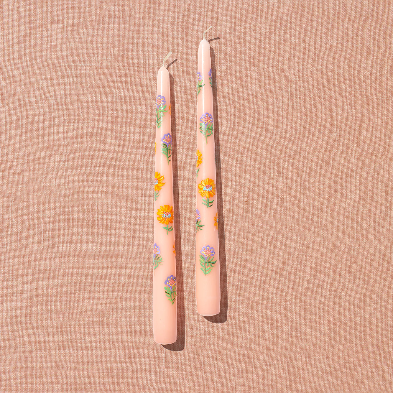 A set of hand painted tapered candles with floral motifs in soft, spring hues such as gold and violet laid out on a peach backdrop.