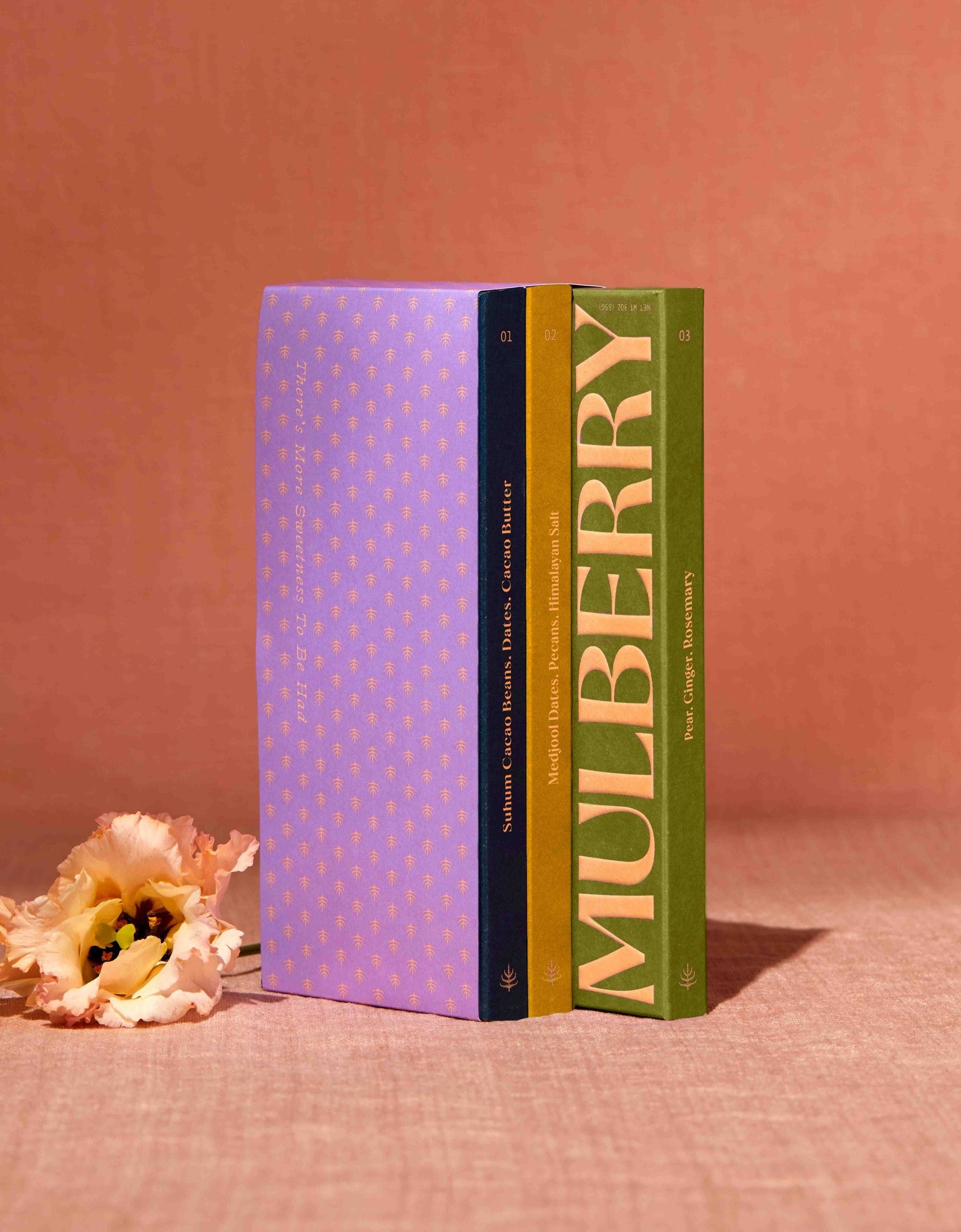 A gift set of 3 date sweetened chocolate bars with no added sugar stands on a peach linen backdrop. To the left of the gift set is a delicate flower. The gift set resembles a library set of books and is wrapped in purple paper adorned with a mulberry motif. The 3 chocolate boxes include: Pure Dark in a navy box; Medjool Date, Pecan, Himalayan Salt in a mustard box; and, Pear, Ginger, Rosemary in a leaf green box.