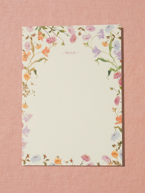 A menu card sits on a peach, linen backdrop. The menu card features a border of handpainted florals by Raleigh-based artist Inslee Fariss. The flowers are inspired by the sunset-colored packaging of Spring & Mulberry chocolate bars.