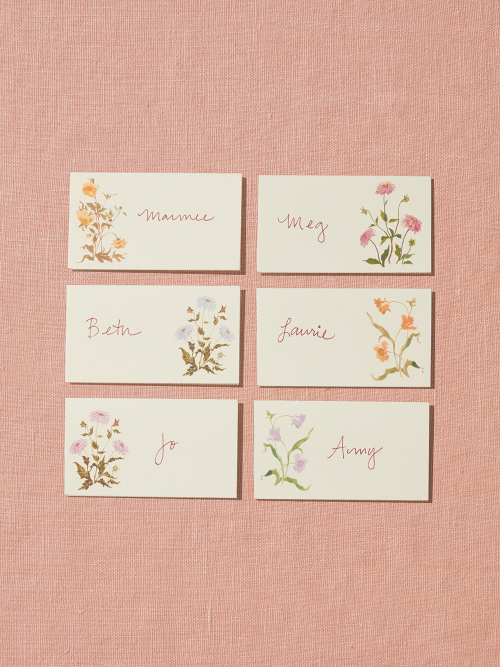 A set of six placecards sit on a peach, linen backdrop. The placecards each feature a different hand-painted flower illustrated by Raleigh-based artist Inslee Fariss. The flowers are inspired by the sunset-colored packaging of Spring & Mulberry chocolate bars.