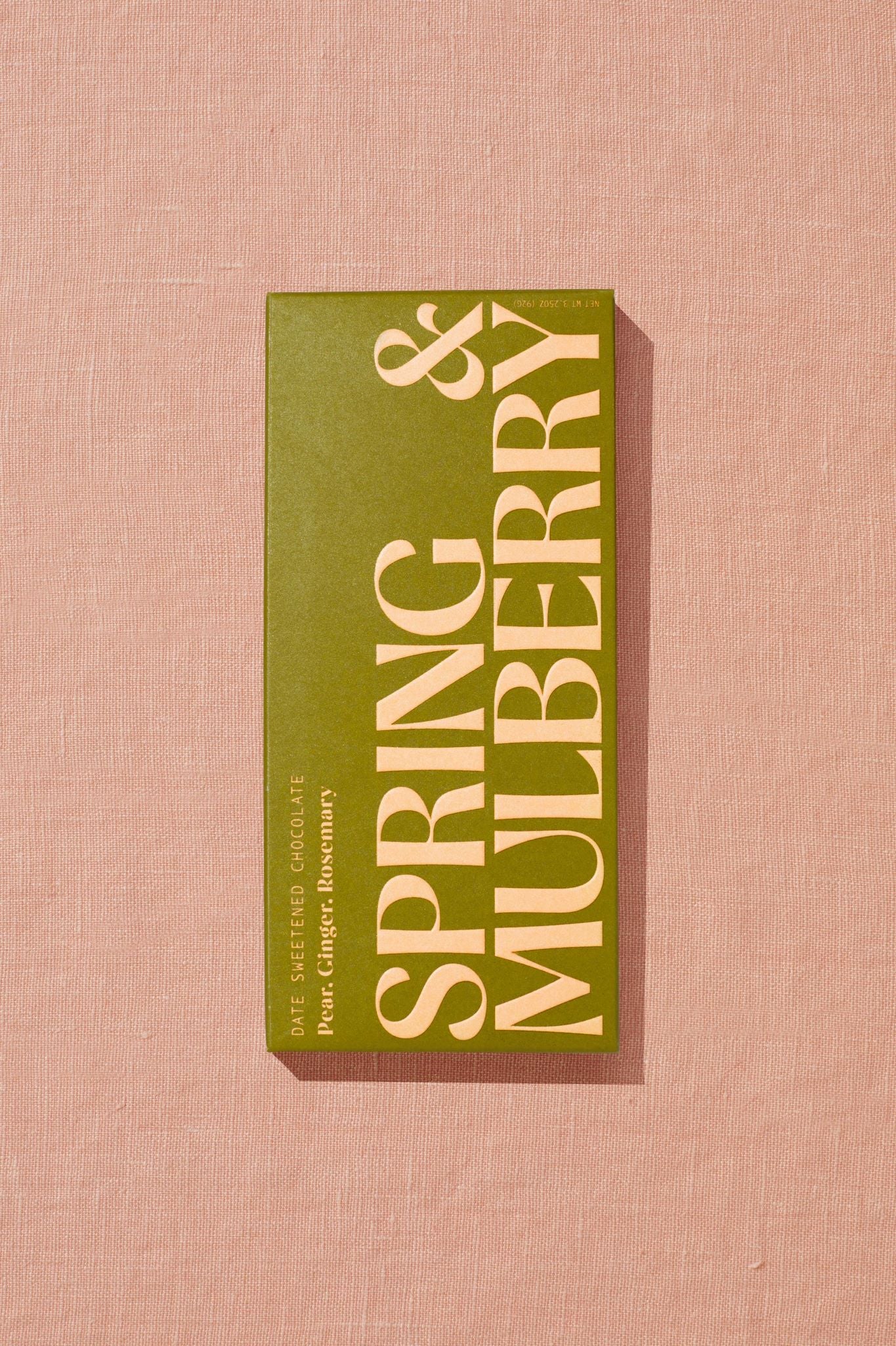 A rectangular, leaf green box containing a 72% date-sweetened dark chocolate bar with no added sugar sits upon peach linen fabric. Warm, direct light hits the box, which reads Date Sweetened Chocolate: Pear, Ginger, Rosemary. Just below sits an oversized, embossed logo that goes edge-to-edge reading Spring & Mulberry in capped font.