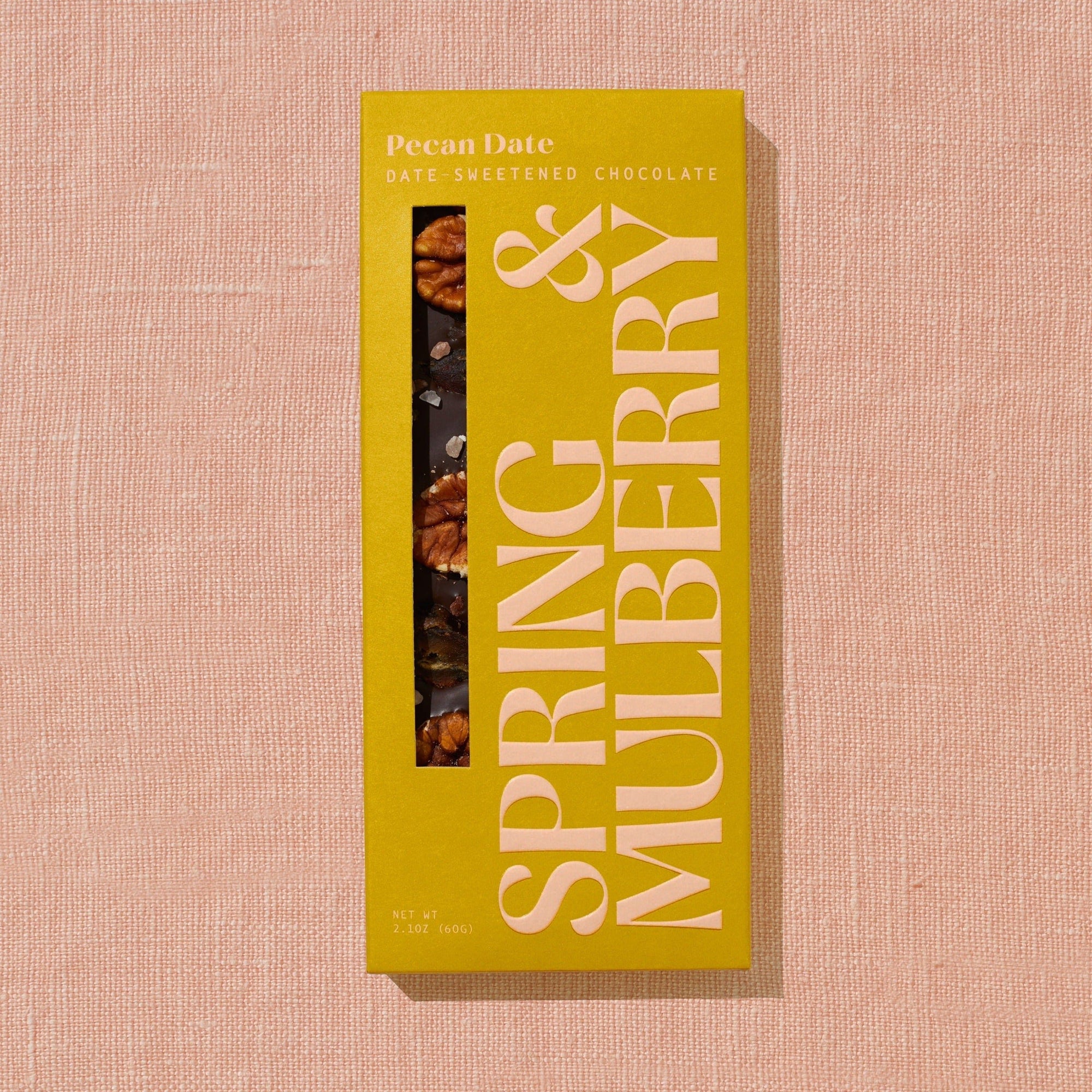 A bright golden Spring & Mulberry box labled Pecan Date date-sweetened chocolate is laid on a warm brown background.