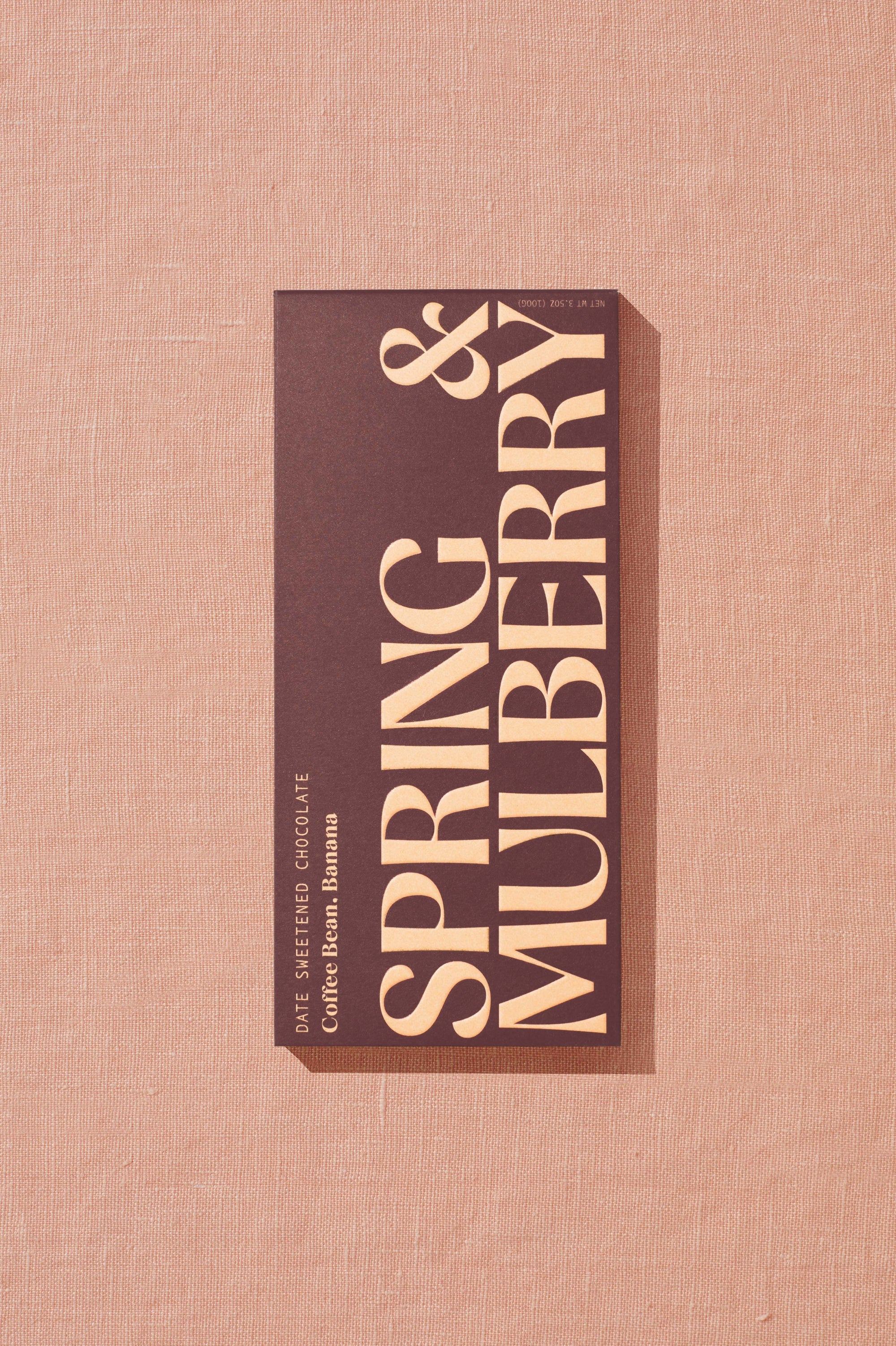 A rectangular, chocolate brown box containing a 72% date-sweetened dark chocolate bar with no added sugar sits upon peach linen fabric. Warm, direct light hits the box, which reads Date Sweetened Chocolate: Coffee Bean, Banana. Just below sits an oversized, embossed logo that goes edge-to-edge reading Spring & Mulberry in capped font.