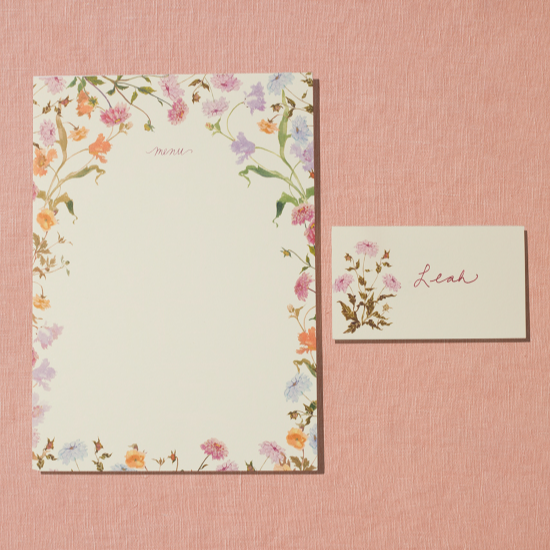 A menu and placecard sit on a linen peach backdrop. The menu card on the left features a border of handpainted florals on a creamy white card. The placecard on the right features one pink flower and a handwritten name.