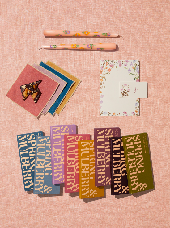 A gift set of 8 date-sweetened chocolate bars, a set of four cocktail napkins, a menu & placecard set, and two hand-painted candles sits on a peach linen backdrop.