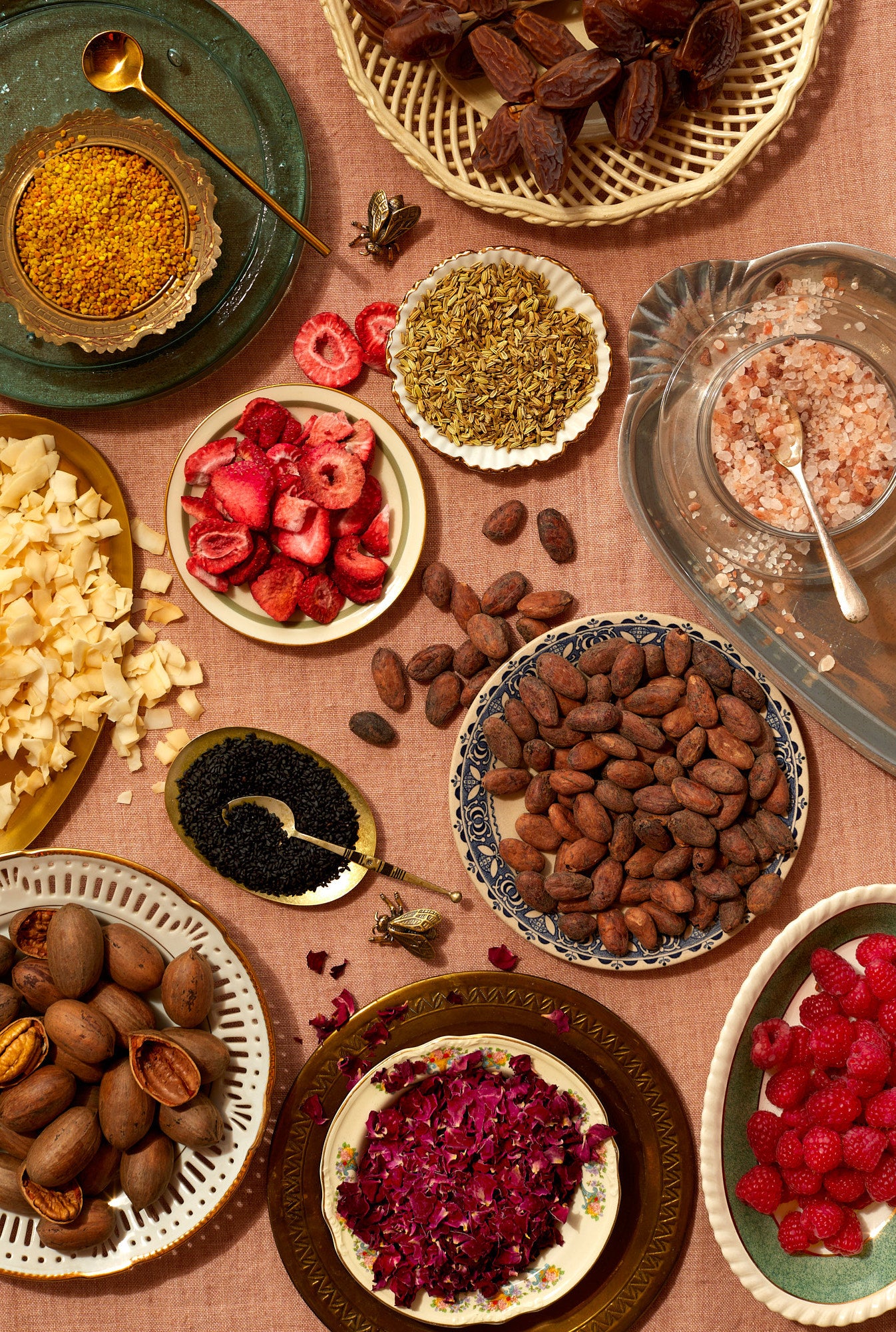 Shot with an overhead view, a mix-and-match assortment of vintage plates, glasses, and bowls filled with flowers, berries, nuts, seeds, dates, and cacao beans fill the image, which is set on a peach linen tablecloth.