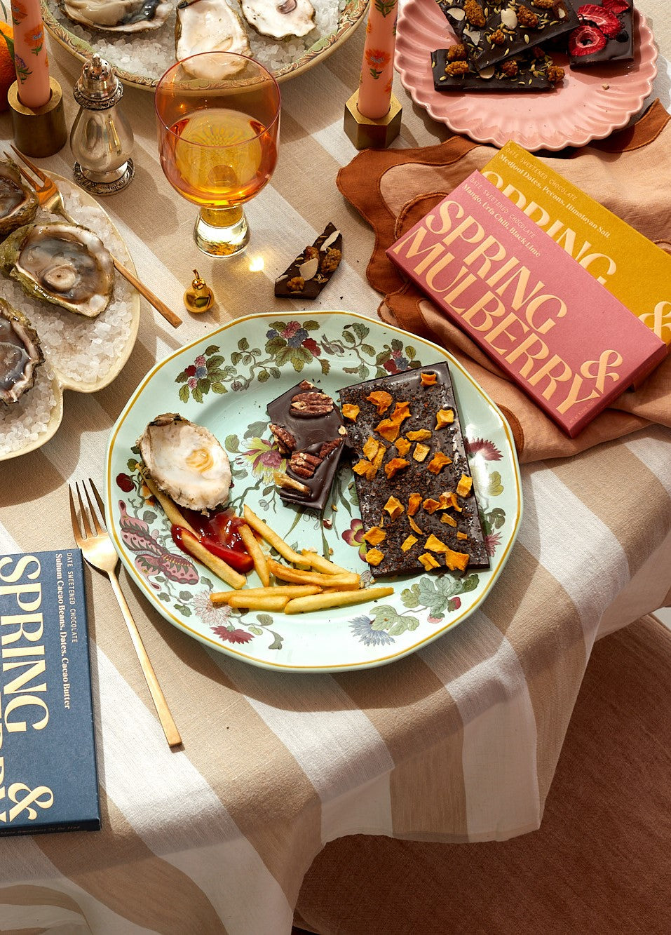 The image shows what remains of a beautiful dinner party where french fries, oyster shells, and Spring & Mulberry date sweetened dark chocolate bars sit upon a plate of turquoise and flower patterned china. Florals, glasses of wine, and plates sit upon a tan and white stripped tablecloth. Spring & Mulberry's Mango, Urfa Chili, Black Lime rose-colored chocolate box and Medjool Date, Pecan, Himalayan Salt mustard box sit in the upper right upon a rose colored napkin with burnt scalloped trim.