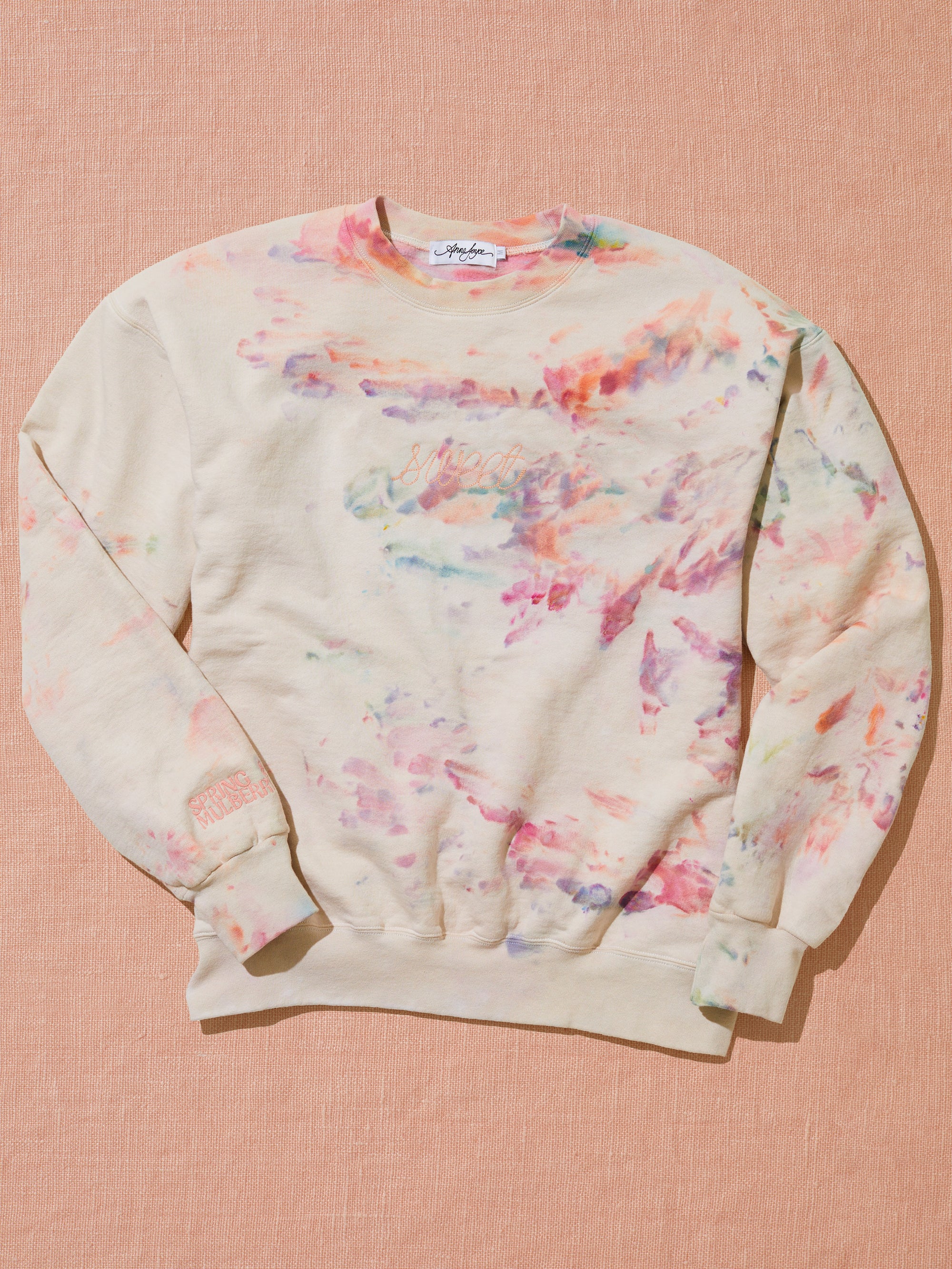 An ice-dyed sweatshirt chainstitched with the word "sweet" sits on a peach fabric backdrop.