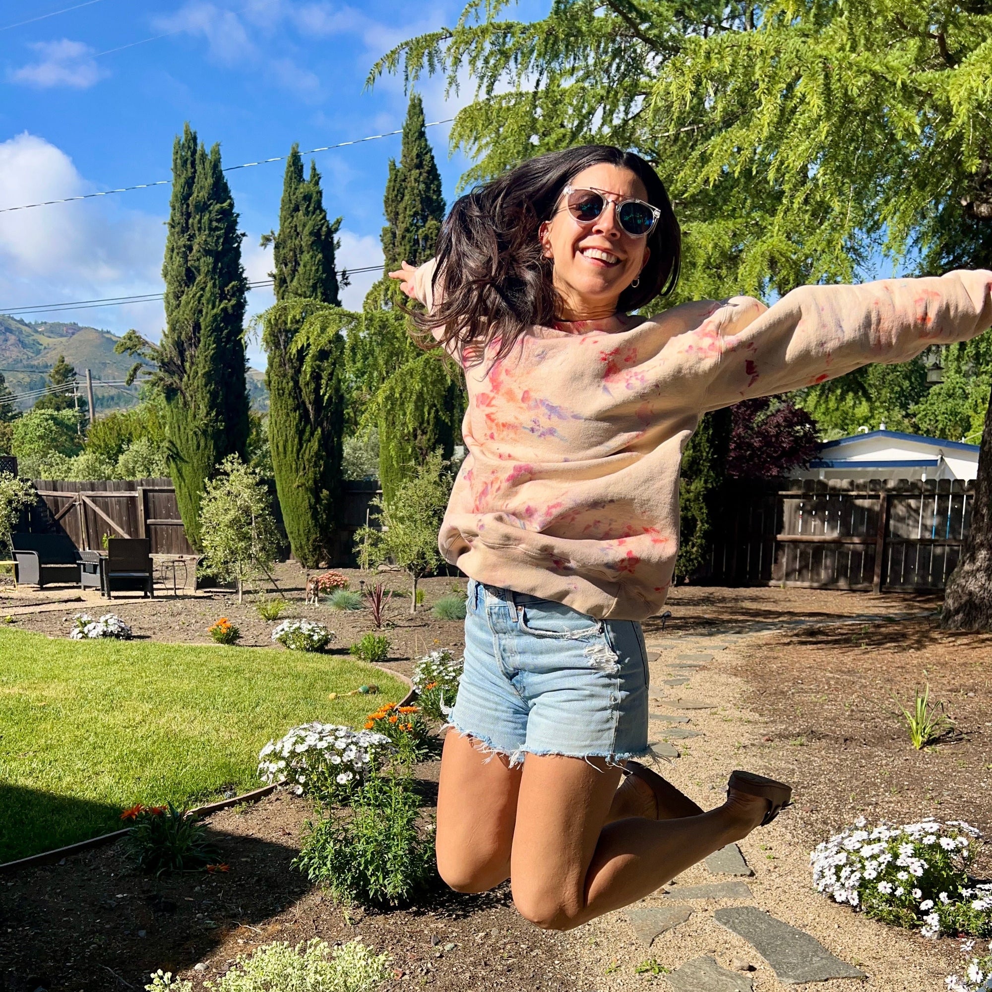 A smiling girl in sunglasses that is wearing the Spring & Mulberry sweatshirt is jumping outside in a garden on a sunny day.