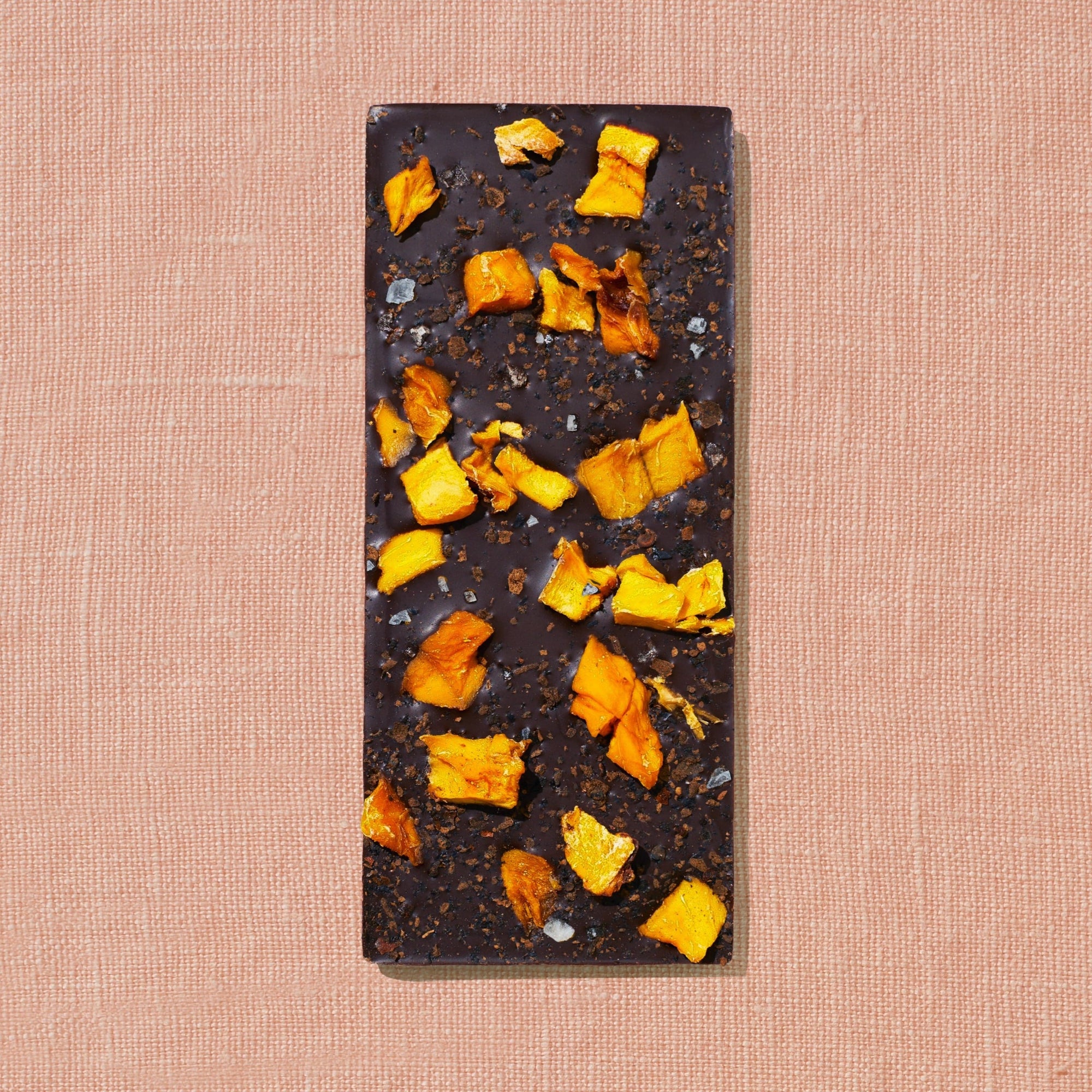 One chocolate bar that is topped with mango pieces, chili flakes, and salt flakes is laid out on a warm brown background.