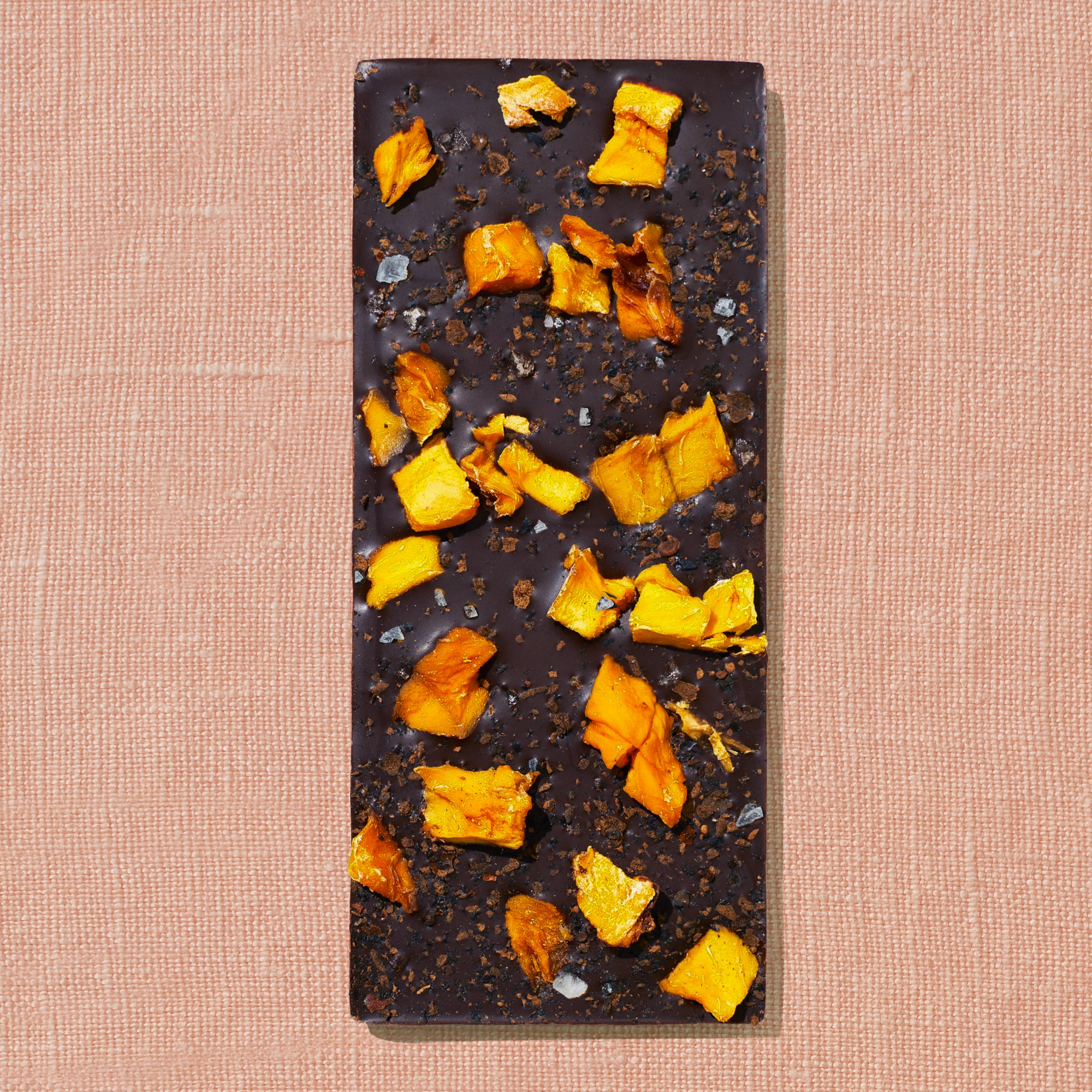 One chocolate bar that is topped with mango pieces, chili flakes, and salt flakes is laid out on a warm brown background.