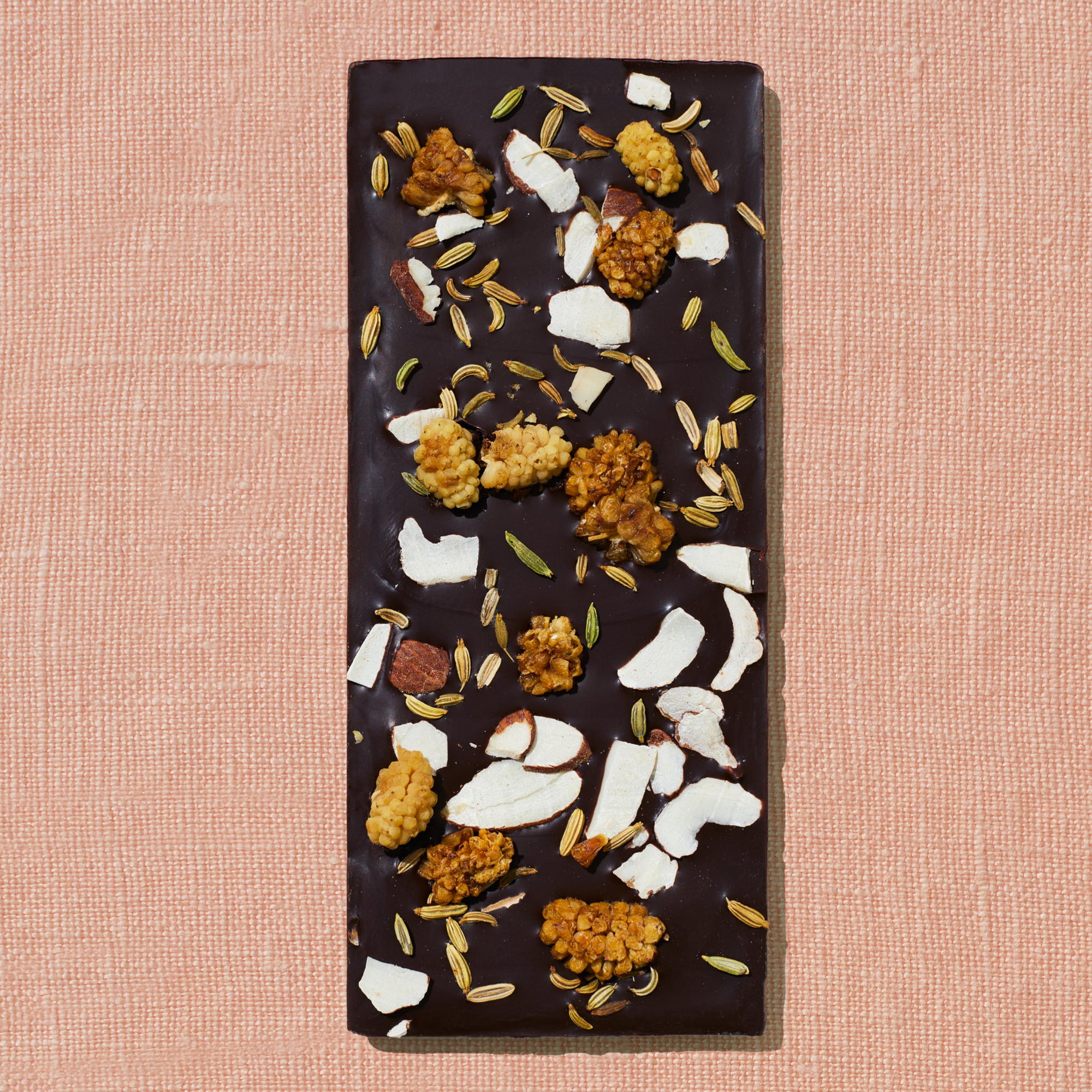 One chocolate bar that is topped with mulberries, almond flakes, and fennel is laid out on a warm brown background.