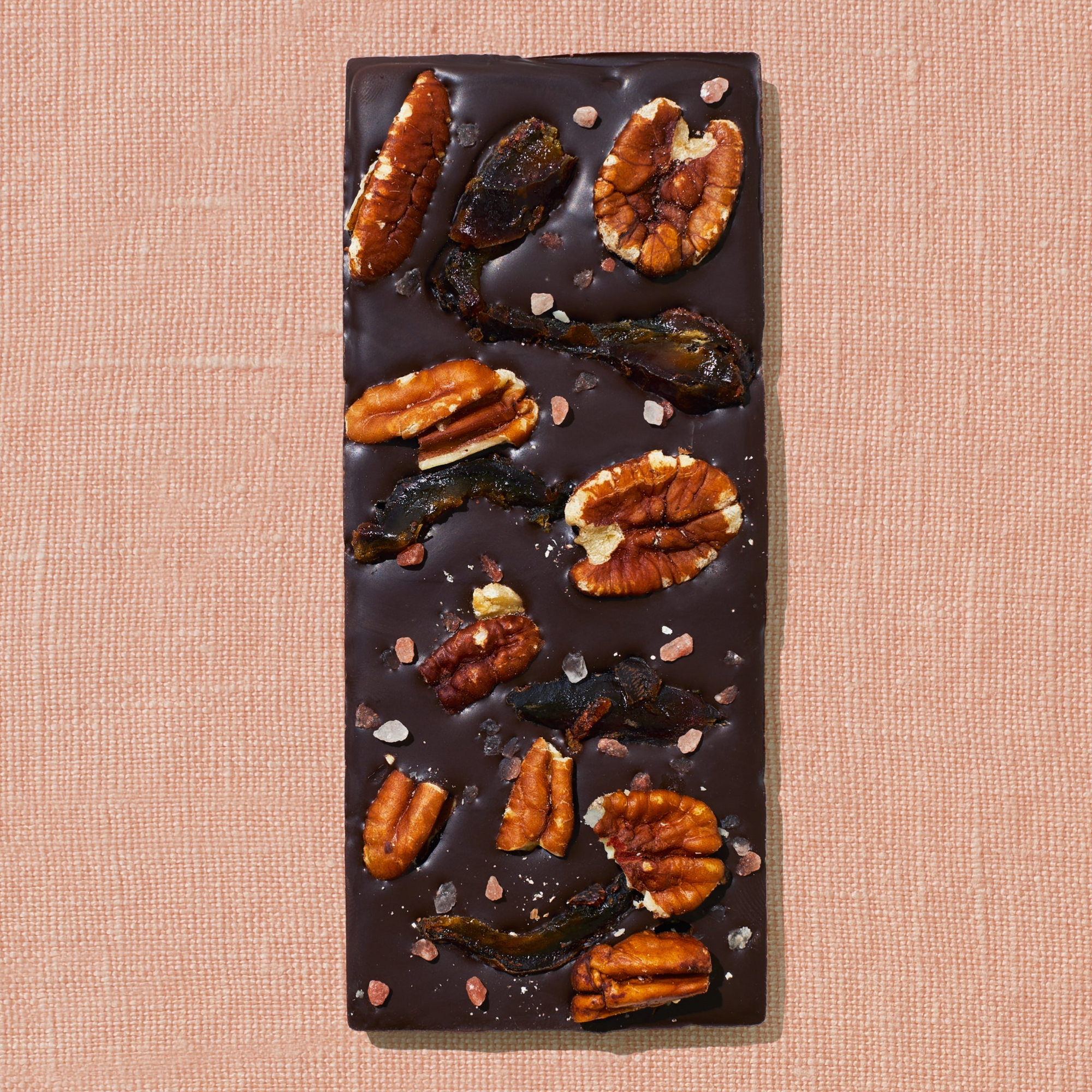 One chocolate bar that is topped with dates, pecans, and rock salt is laid out on a warm brown background.