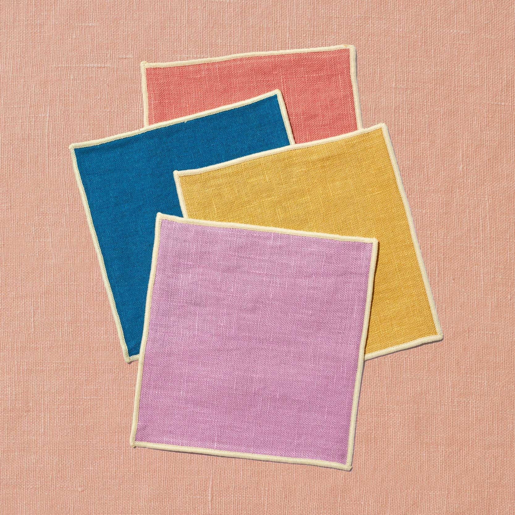 A set of four dessert napkins sits upon a peach linen background. The napkins are artfully scattered with rose in the back, followed by marine blue, mustard, and lavender in front. Each napkin matches the colors of Spring & Mulberry's date sweetened chocolate boxes, which are inspired by the setting sun.