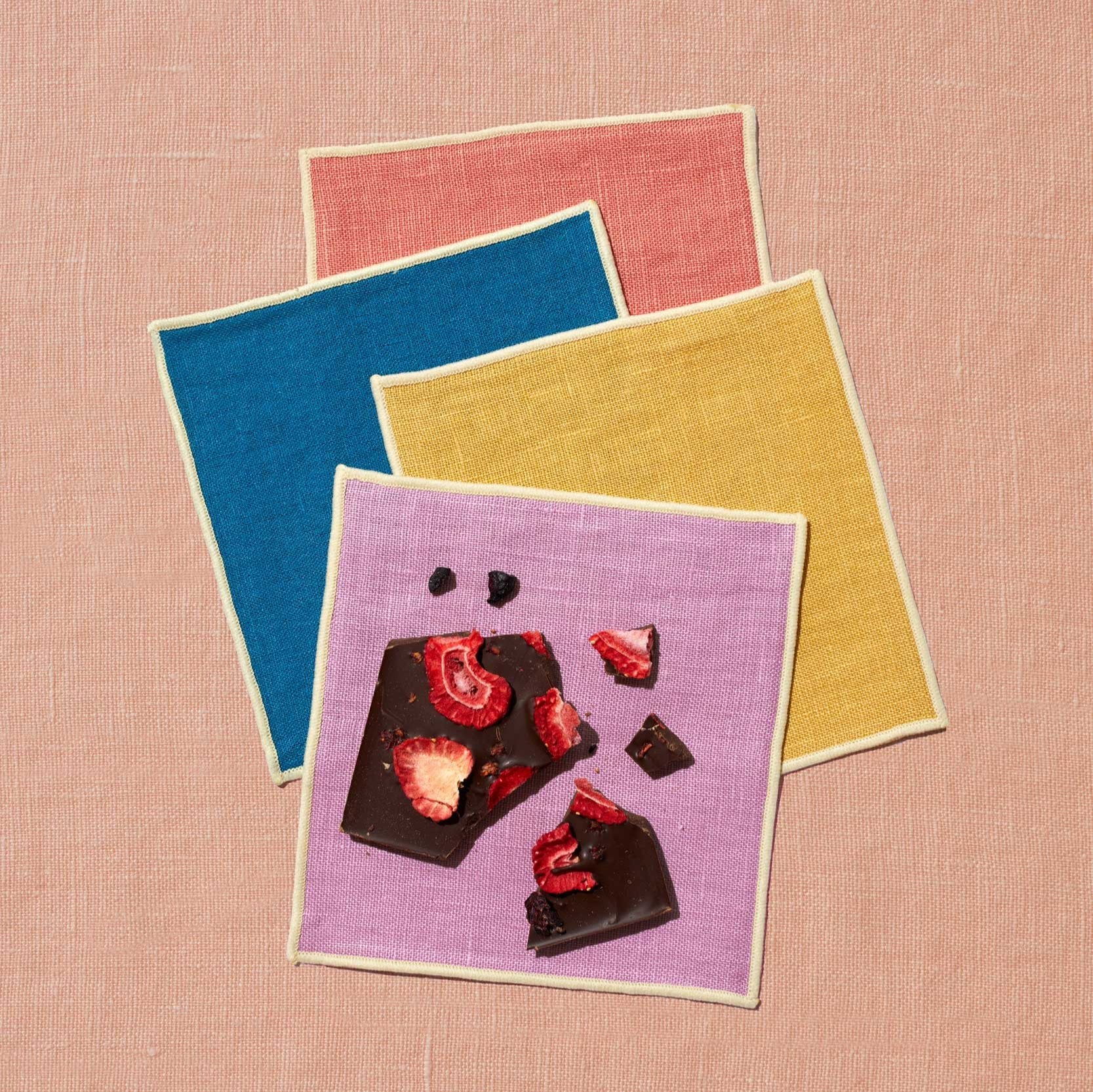 A set of four napkins sits upon a peach linen background. The napkins are artfully scattered and come in four colors with ivory trim: rose, marine blue, mustard, and lavender. On the front napkin, which is lavender, sits a broken bar of date sweetened chocolate topped with mixed berries. The chocolate has no added sugar.