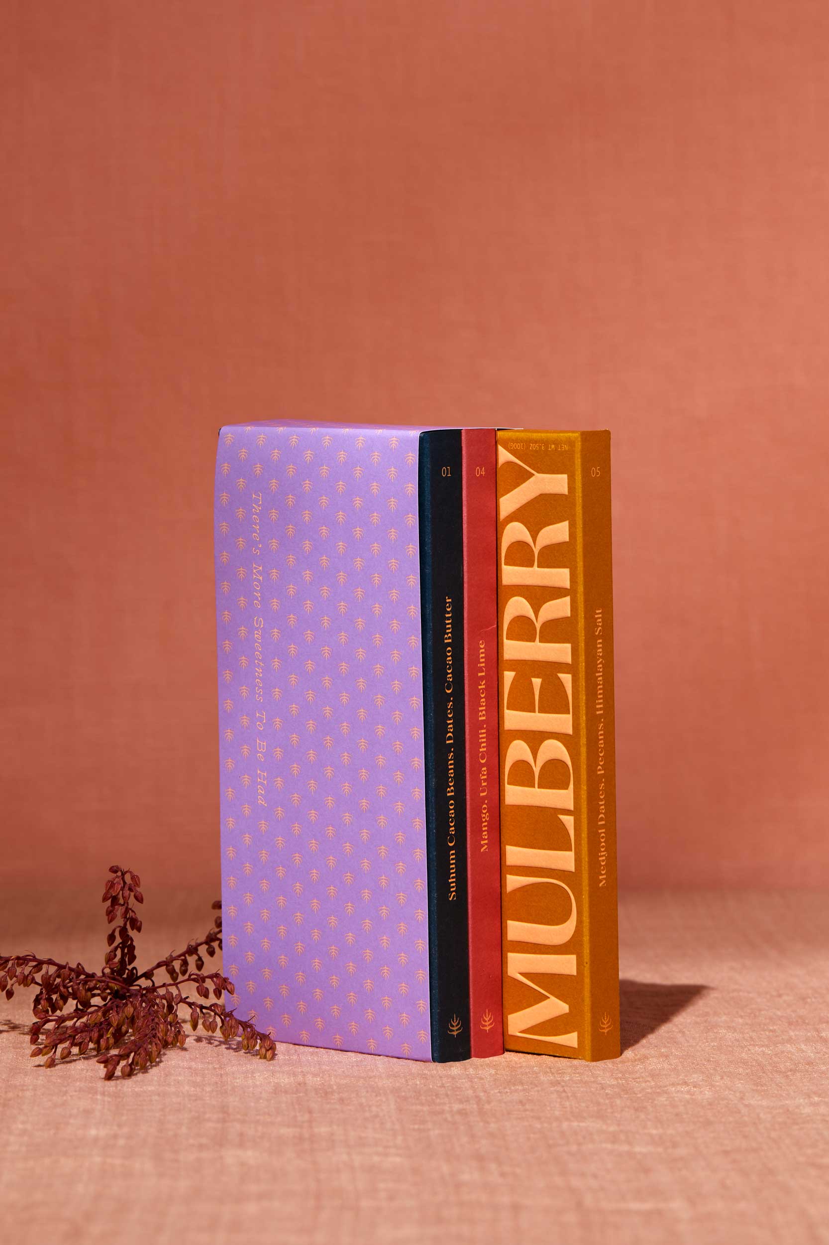 A gift set of 3 date sweetened chocolate bars with no added sugar stands on a peach linen backdrop. To the left of the gift set is a delicate flower. The gift set resembles a library set of books and is wrapped in purple paper adorned with a mulberry motif. The 3 chocolate boxes include: Pure Dark in a navy box; Mango, Urfa Chili, Black Lime in a rose colored box; and, Medjool Date, Pecan, Himalayan Salt in a mustard box.