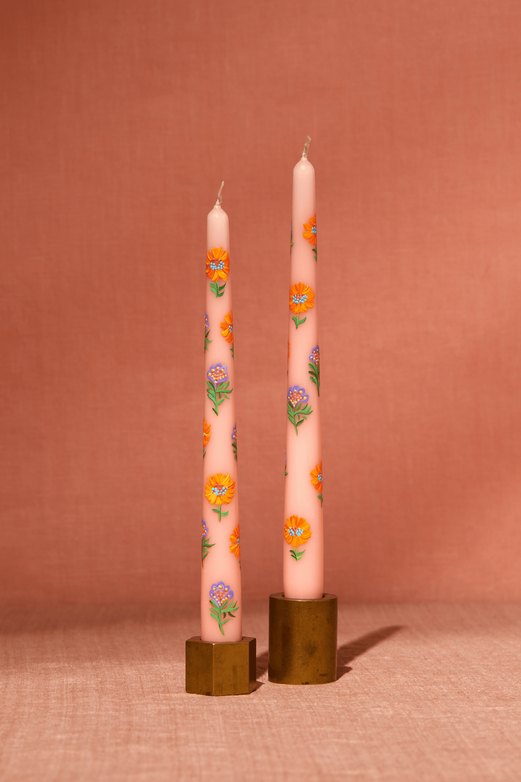 A set of two 10" tapered peach candles stands in brass candlesticks. Each candle is adored with handpainted flowers in peach and lavender shades. The image is shot straight on with warm light on a peach backdrop.