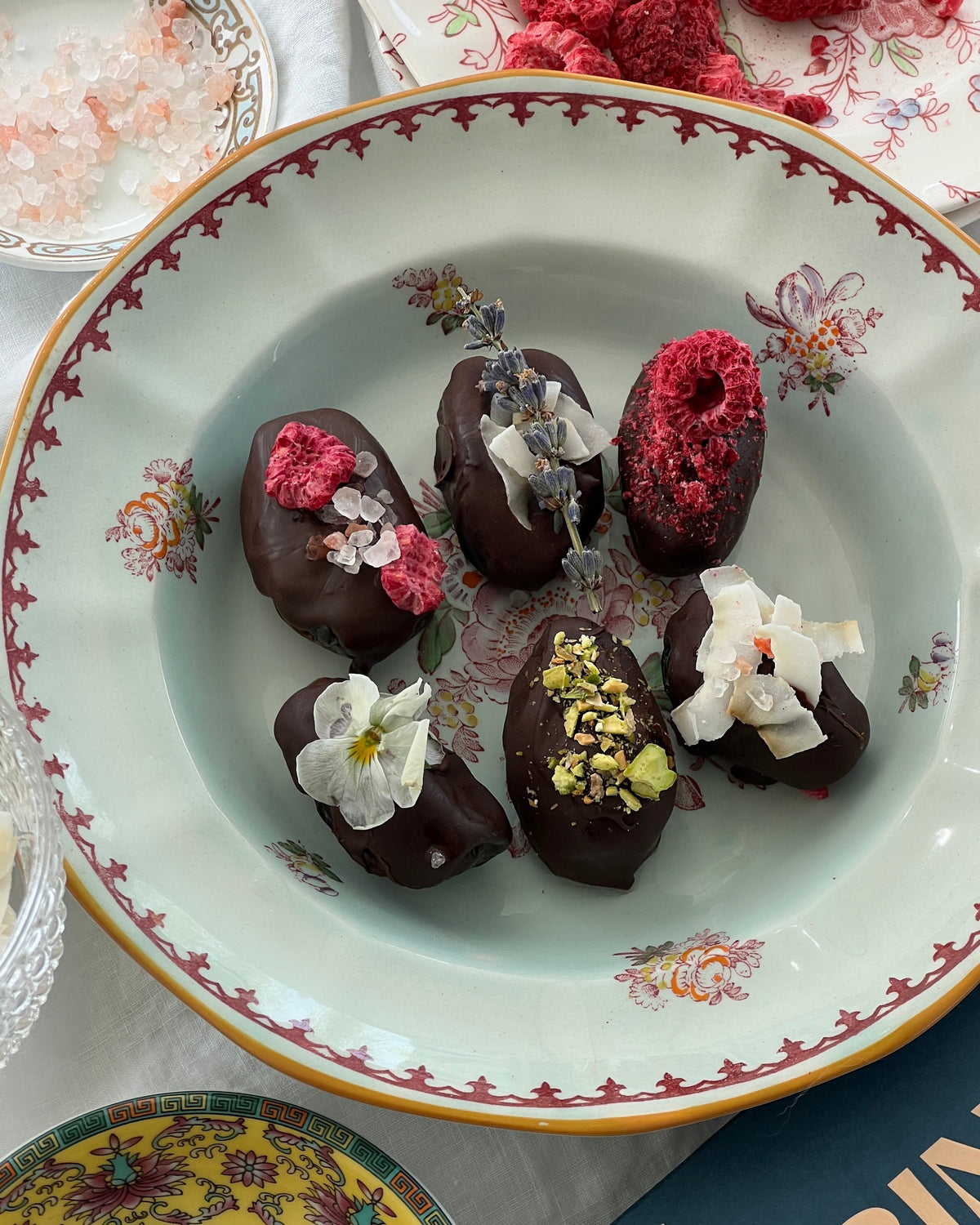 Photographed are six dates covered in dark chocolate, with florals and dried fruits to top. The dates sit in a blue and pink bowl with floral designs printed.
