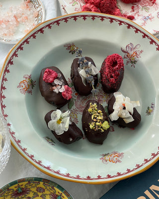 Photographed are six dates covered in dark chocolate, with florals and dried fruits to top. The dates sit in a blue and pink bowl with floral designs printed.
