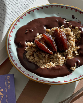 In this photo there is a bowl of oatmeal, with dark chocolate drizzled on top. The bowl sits on a blue and green striped table cloth, next to an open book, and a Spring & Mulberry Earl Grey bar in a purple box. 