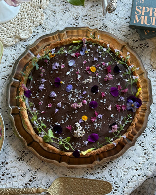 Pictured is a chocolate tart with purple, pink and green florals on top. The tart is in a silver dish, on top of a white lace tablecloth. In the corner is a Spring & Mulberry Sea Salt chocolate bar, and silverware for serving.