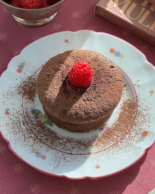 Pictured is a small chocolate cake, topped with a raspberry and cacao dust. Served on a floral glass plate, and a pink tablecloth. 