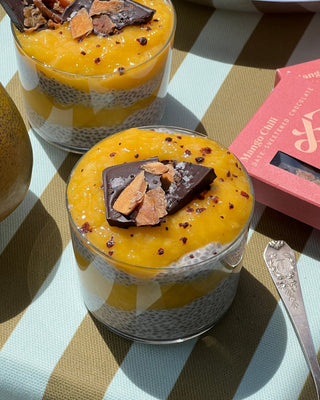 Photographed is a cup layered with mango, chocolate, and chia seed pudding, set atop a blue-and-green striped tablecloth. Beside the cup sits a silver spoon and a Spring & Mulberry Mango Chili chocolate bar.