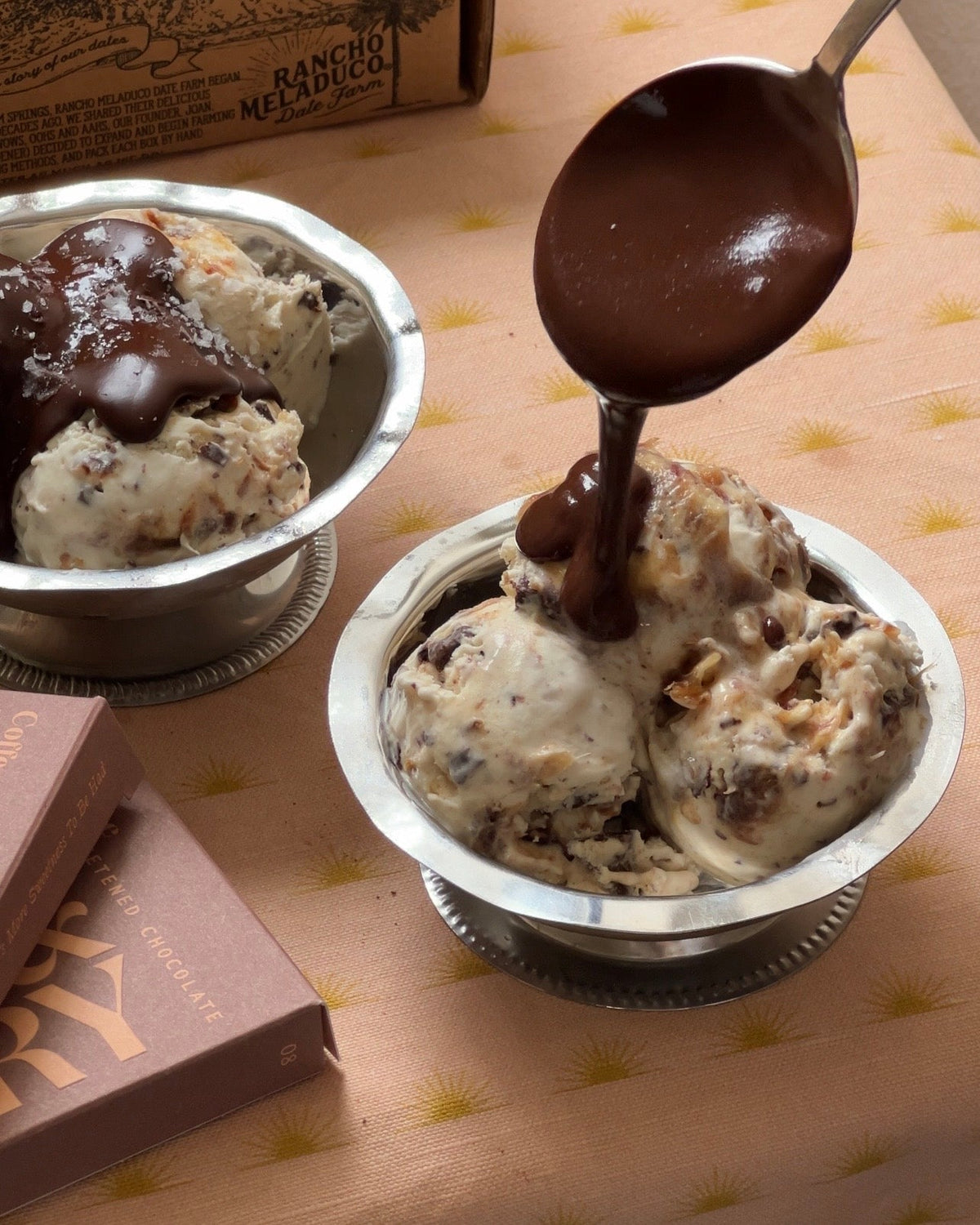 Pictured above is a scoop of ice cream served in a silver cup, over a peach tablecloth. Above the ice cream, there is a spoon dripping melted chocolate atop the ice cream. In the corner, there are two Spring & Mulberry Coffee bars.  