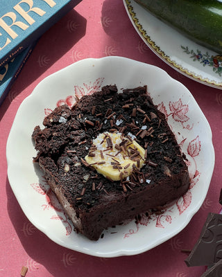 Photographed is a slice of chocolate zucchini bread, with butter and chocolate shavings on top. The bread slice sits on a white plate with red florals, and a maroon tablecloth. 