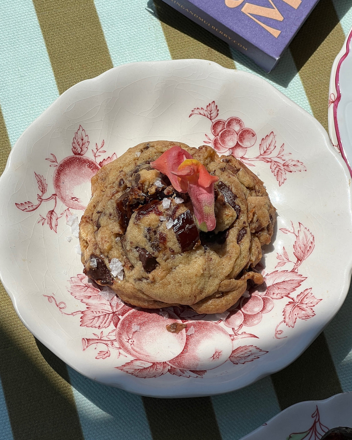 Photographed is a chocolate chip cookie, served on a red and white floral plate. The plate sits on a green and blue stripe tablecloth, with a purple Spring & Mulberry bar in the top right corner.  