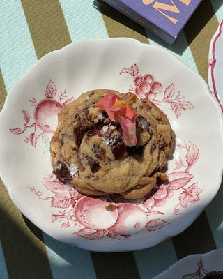 Photographed is a chocolate chip cookie, served on a red and white floral plate. The plate sits on a green and blue stripe tablecloth, with a purple Spring & Mulberry bar in the top right corner.  