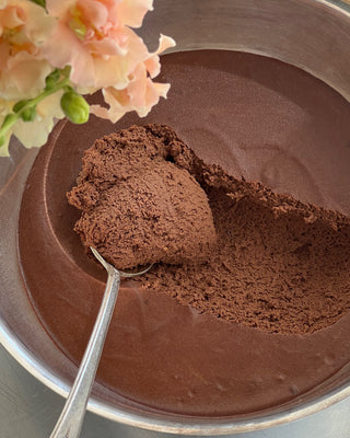 Photographed is a large bowl of chocolate mousse being scooped with a silver spoon. In the upper left corner, there are light pink flowers.
