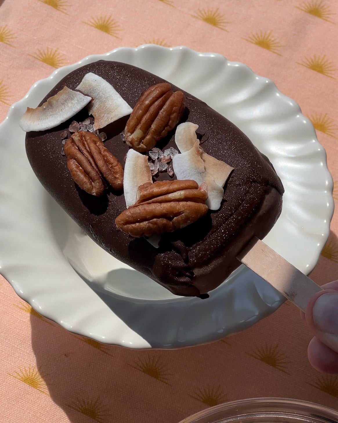 Photographed is a single popsicle covered in dark chocolate, pecans, and coconut chips. The popsicle sits on a white bowl, with ruffle edges, and a peach tablecloth. 