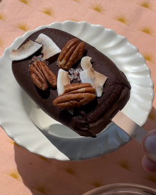 Photographed is a single popsicle covered in dark chocolate, pecans, and coconut chips. The popsicle sits on a white bowl, with ruffle edges, and a peach tablecloth. 