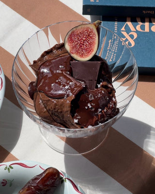 Photographed is a chocolate pudding, topped with sea salt and a fig, in a glass bowl. Under the glass bowl is a brown and blue striped tablecloth, and a Pure Dark Spring & Mulberry bar. 
