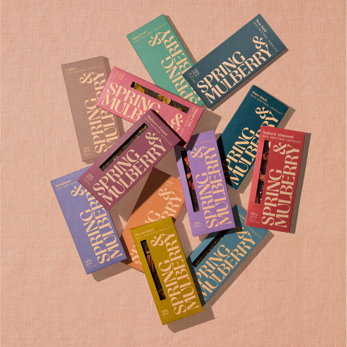 Spring & Mulberry dark chocolate bars in colorful packaging, including Pure Dark, Mint Leaf, Coffee, Mixed Berry, Salted Almond, and other flavors, arranged in an overlapping flat lay against a textured peach background.