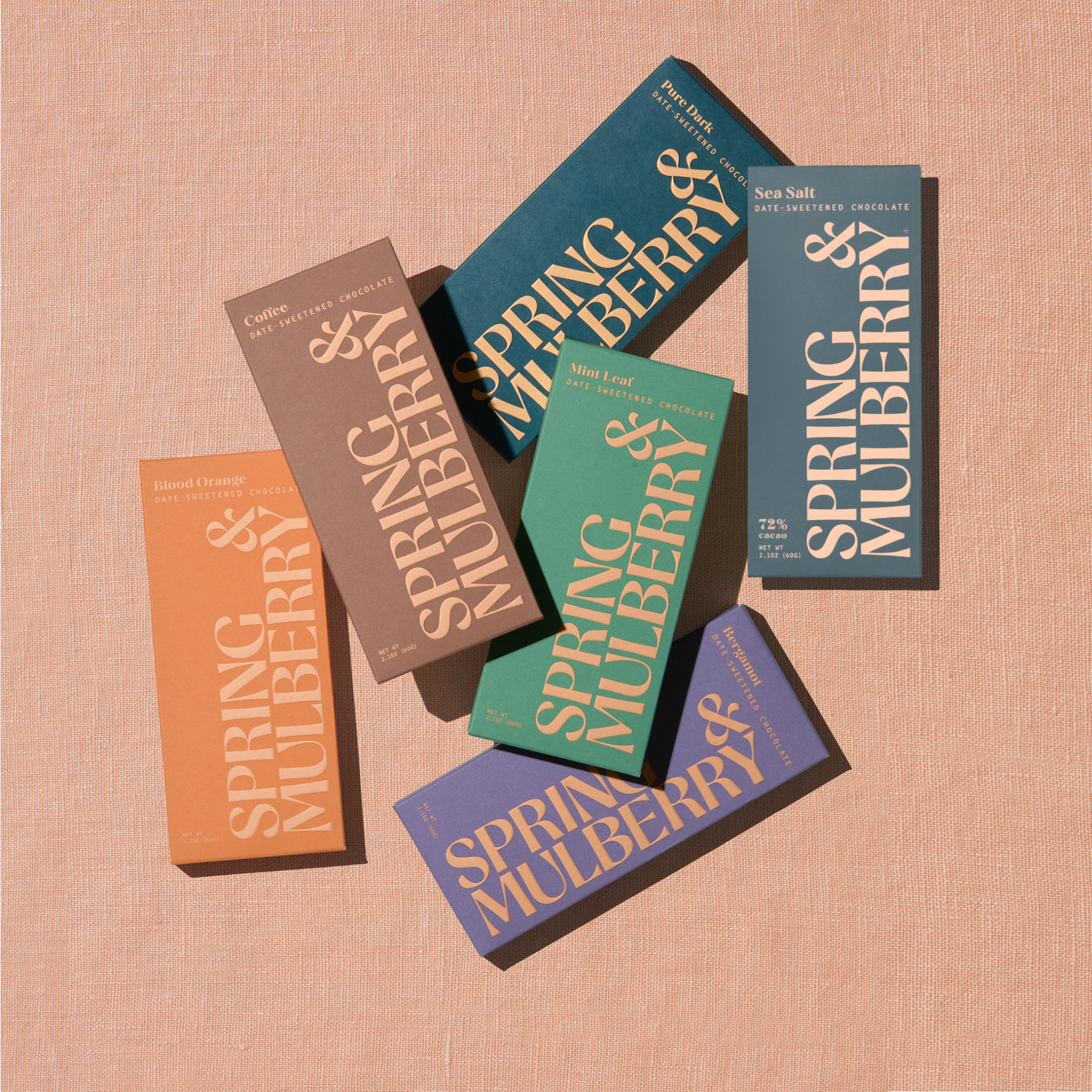 Five artisanal chocolate bars in their Spring & Mulberry boxes are laid out on a surface, each with different color packaging representing various flavors.