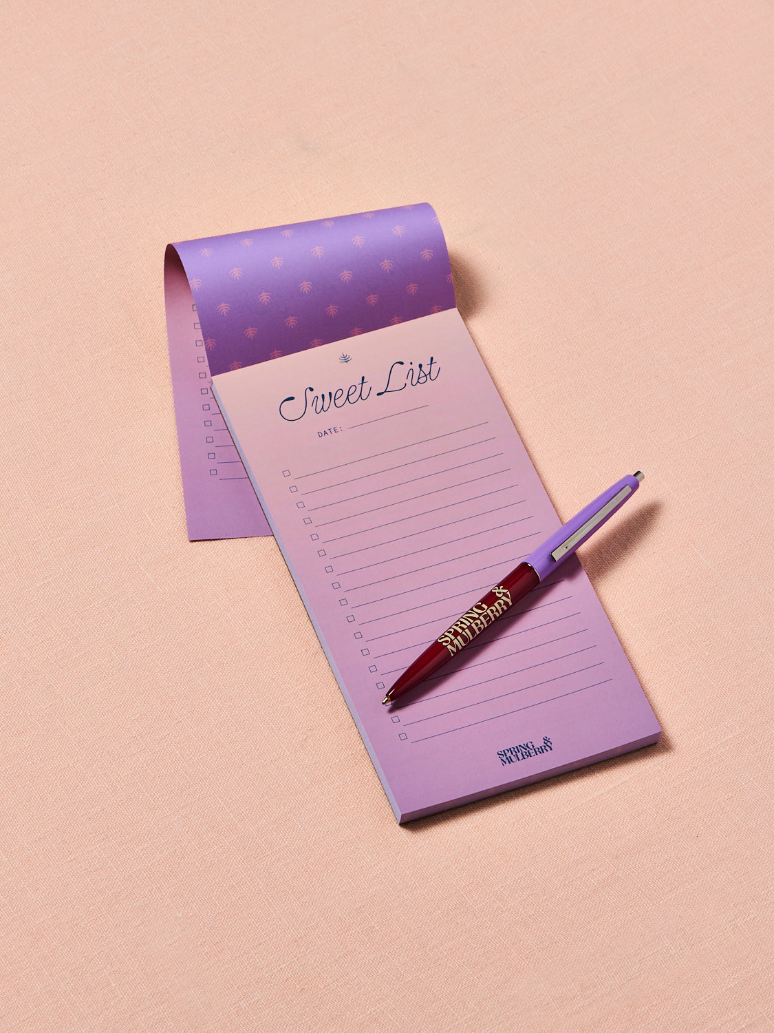 Spring & Mulberry "Sweet List" notepad in soft lavender with a matching branded pen, featuring a checklist layout for notes, errands, and grocery lists on a blush pink background.