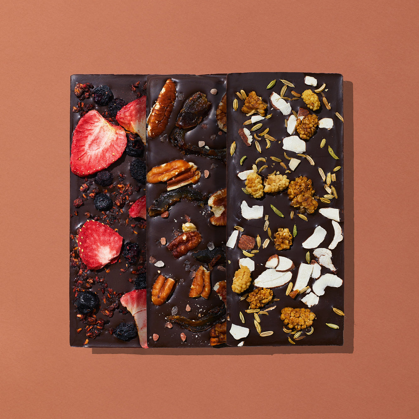 A set of three artisanal chocolate bars layed out flat on a warm brown backgroun, each displaying their toppings. The bars include Mixed Berry, Pecan Date, and Mulberry Fennel.