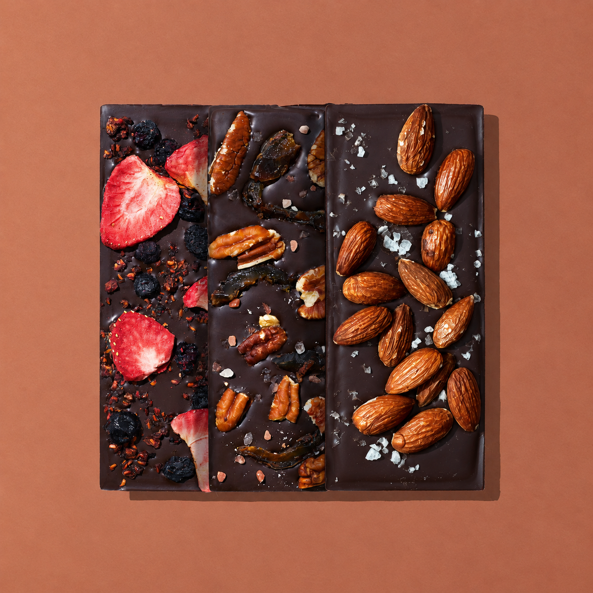 Three Spring & Mulberry dark chocolate bars topped with mixed berries and strawberries, pecans and dates, and whole almonds with flaky sea salt, arranged side by side on a warm terracotta background.
