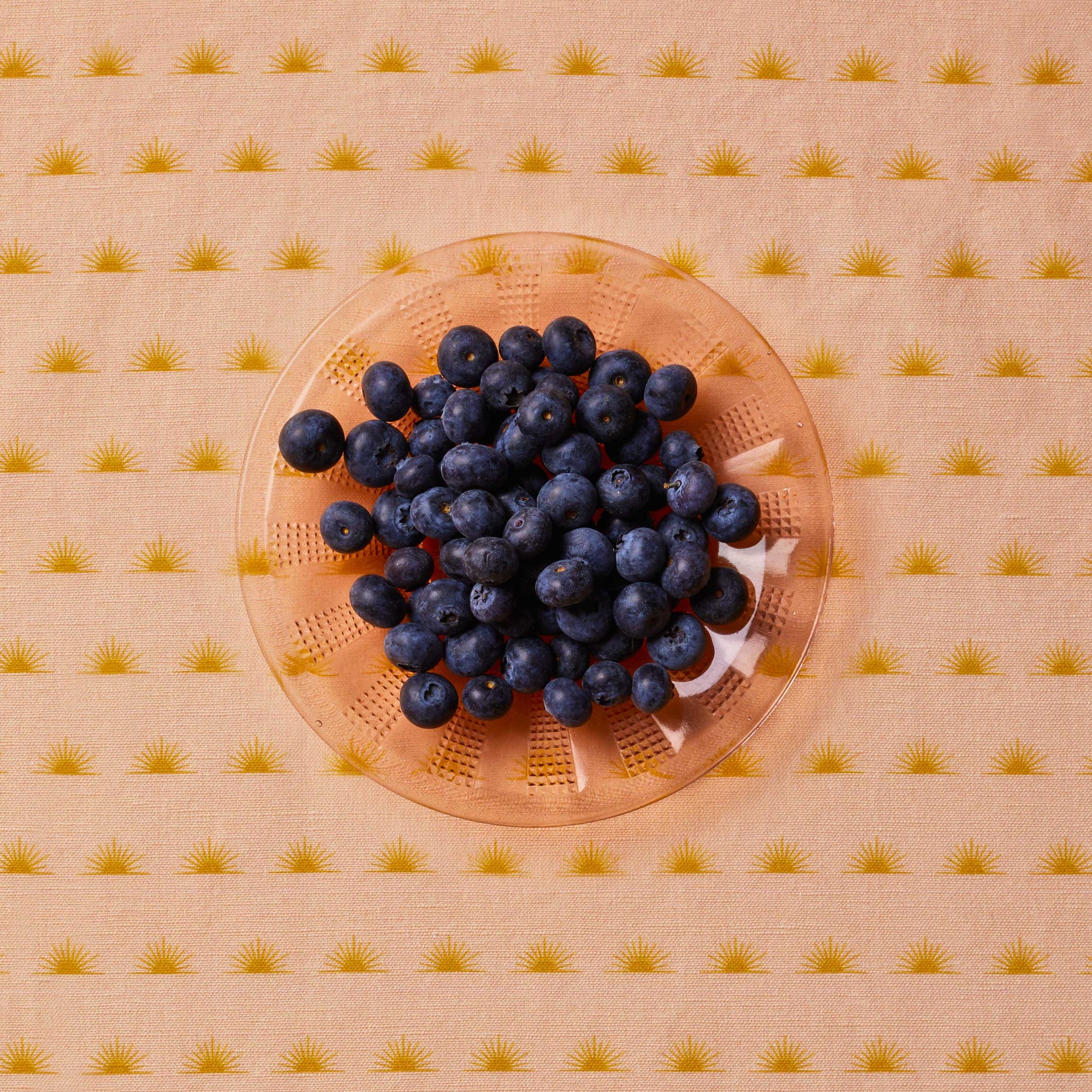 Blueberries sitting in a peach-colored glass bowl on top of a patterned tablecloth