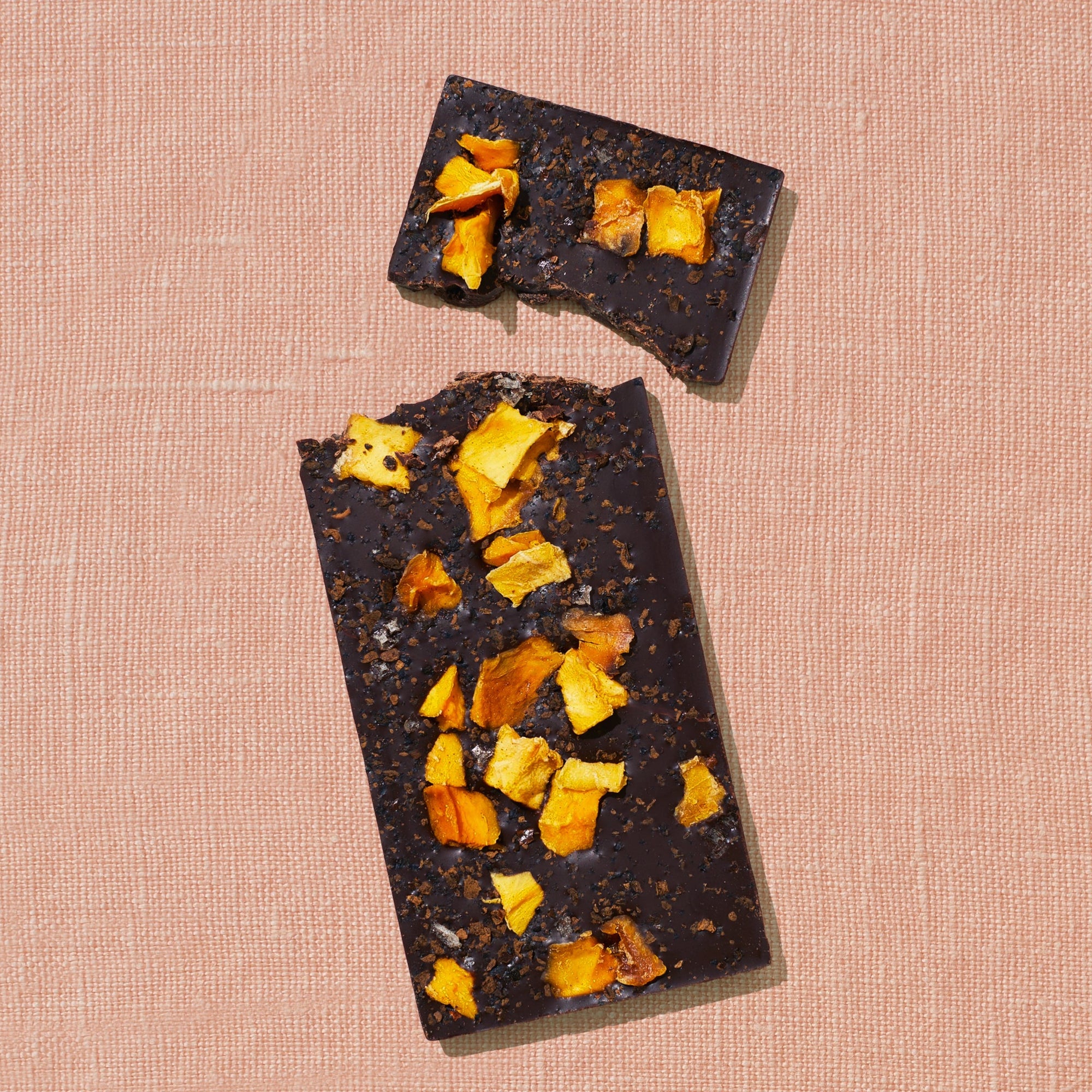 One chocolate bar that is topped with mango pieces, chili flakes, and salt flakes is laid out on a warm brown background broken in two pieces.